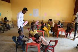 A teacher standing among young children seated at small tables in a reopened classroom.