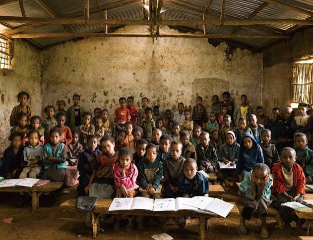 A crowded classroom of primary school students seated at wooden benches with open exercise books.